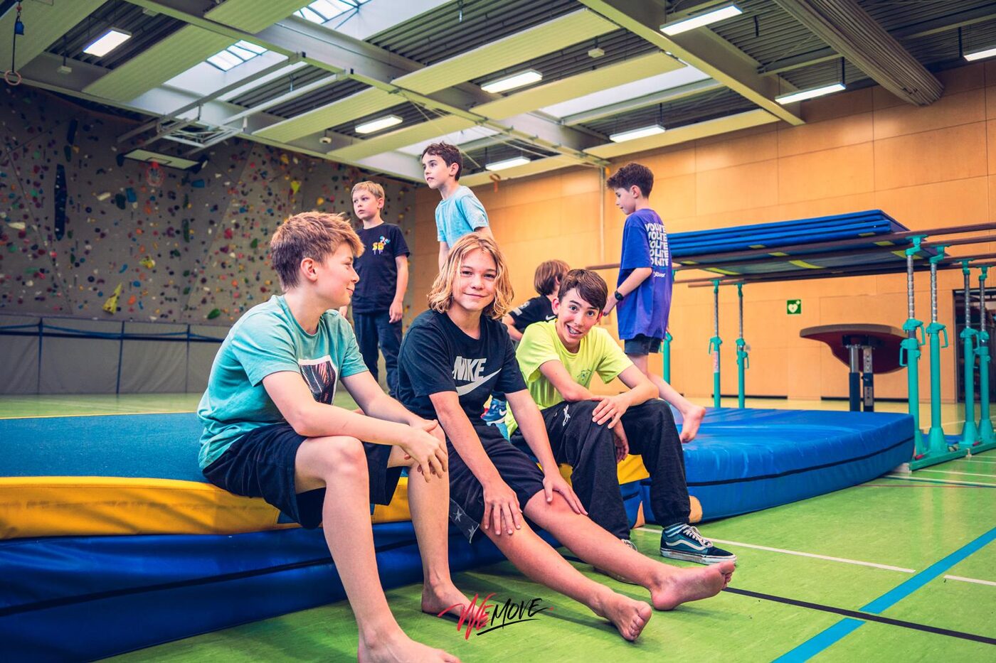 Jugendliche sitzen auf Weichböden in einer Sporthalle, im Hintergrund Parkour-Aufbauten